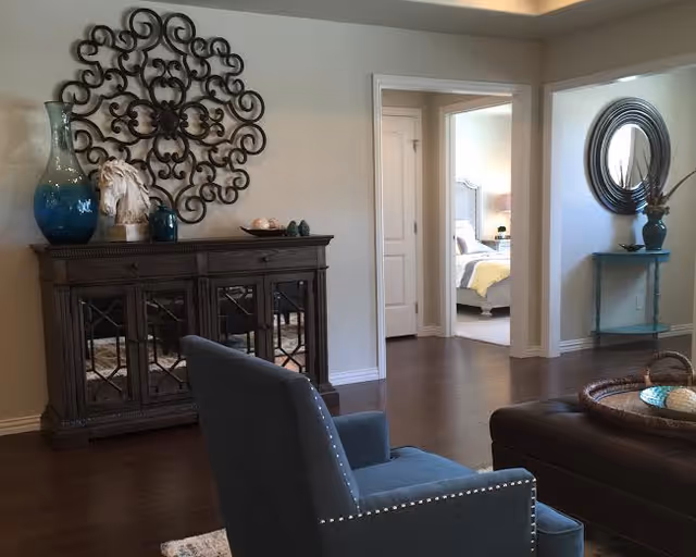 Interior view of a living room area with a dark wooden cabinet adorned with decorative items including a large blue vase and a white horse sculpture. A blue upholstered chair with nailhead trim is in the foreground, and a brown leather ottoman with a woven tray is partially visible. The room has hardwood floors and beige walls. Two doorways lead to a bedroom with a bed and nightstand visible, and a small blue table with a round mirror above it is against the wall near the second doorway.