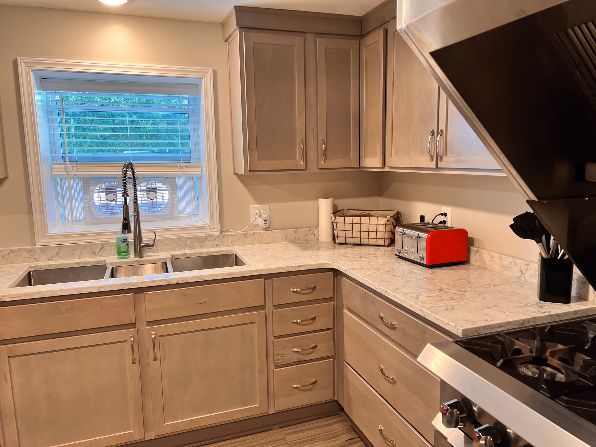 Modern kitchen with marble-look countertops, a double sink beneath a window, light wood cabinets, a red toaster, and a stainless steel range.