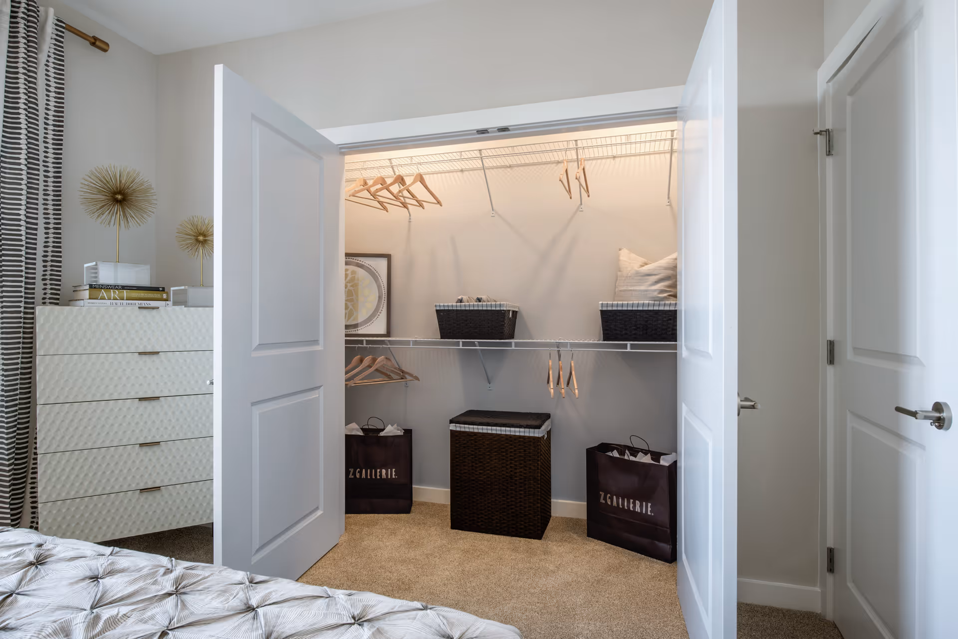 Open closet with white double doors in a bedroom. Inside the closet are wooden hangers on white wire shelves, two black baskets, a framed picture, a black laundry hamper, and two black shopping bags labeled 'Z GALLERIE'. To the left of the closet is a white dresser with books and decorative gold sculptures on top. The room has beige carpet and light-colored walls.
