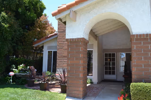 Front entrance of a single-story building with an arched porch supported by brick columns, potted plants, and a grassy lawn.