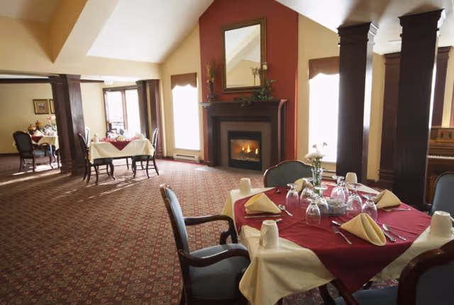 A cozy dining room with tables set for a meal, featuring white tablecloths with red overlays, napkins, glassware, and silverware. The room has a patterned carpet, large windows with curtains, a fireplace with a mirror above it, and dark wooden columns and furniture.