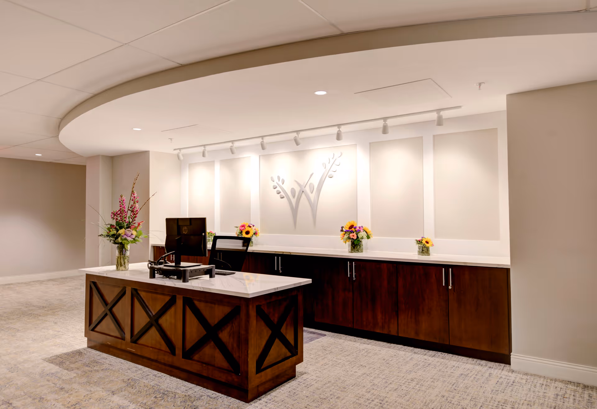 Reception desk area with a dark wood front and white marble countertop, decorated with vases of colorful flowers. Behind the desk is a long cabinet with dark wood doors and a white countertop, above which is a wall with a decorative emblem and track lighting. The space has a neutral color palette with beige walls and carpeted floor.