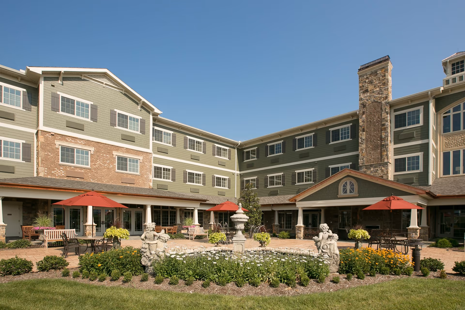 Outdoor courtyard area of a senior living facility with a central fountain surrounded by flowers and greenery. There are several benches and tables with red umbrellas on a paved patio, and the building has green siding with multiple windows and a stone chimney under a clear blue sky.