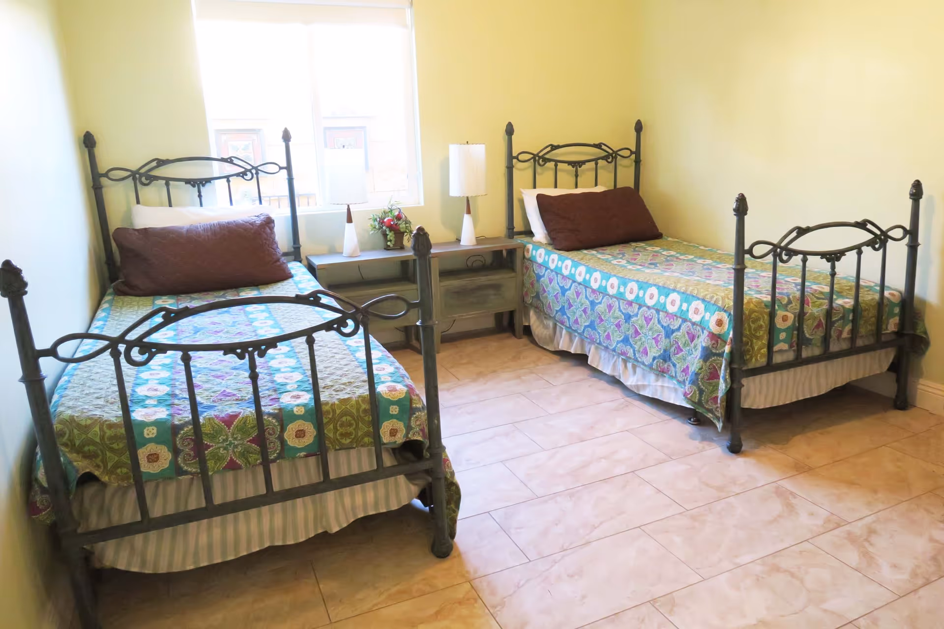 A small bedroom with two single beds featuring black metal frames and colorful patterned bedspreads. Between the beds is a wooden nightstand with two white lamps and a small flower arrangement. A window behind the nightstand lets in natural light, and the floor is tiled.
