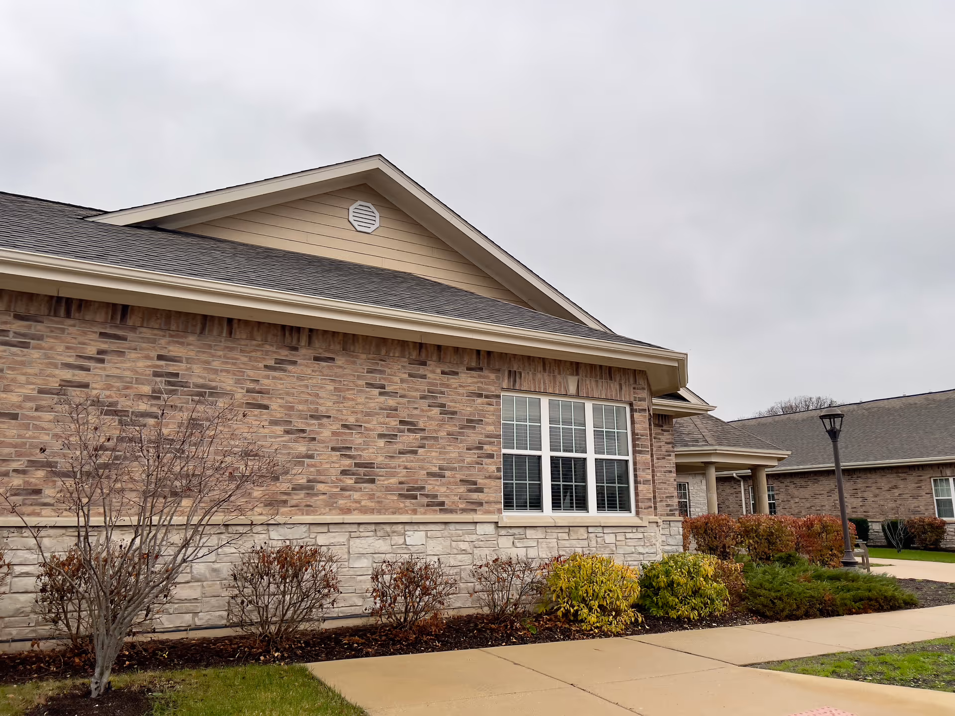 Exterior view of a single-story brick-and-stone senior living building with landscaped shrubs and a sidewalk under an overcast sky.