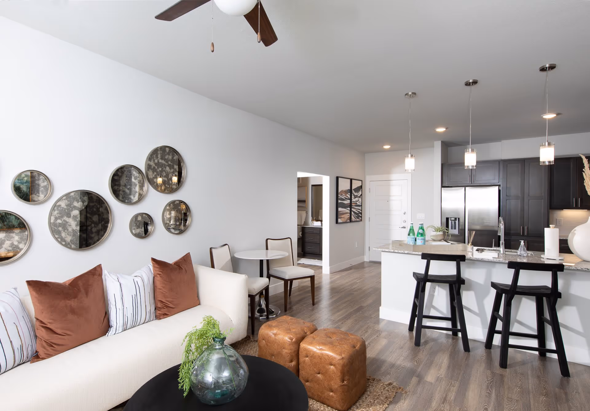A modern living room featuring a white sofa with brown and striped pillows, a round black coffee table, and decorative mirrors on the wall. A small dining area with a round table and chairs is visible, along with a kitchen bar area equipped with stools and modern appliances.