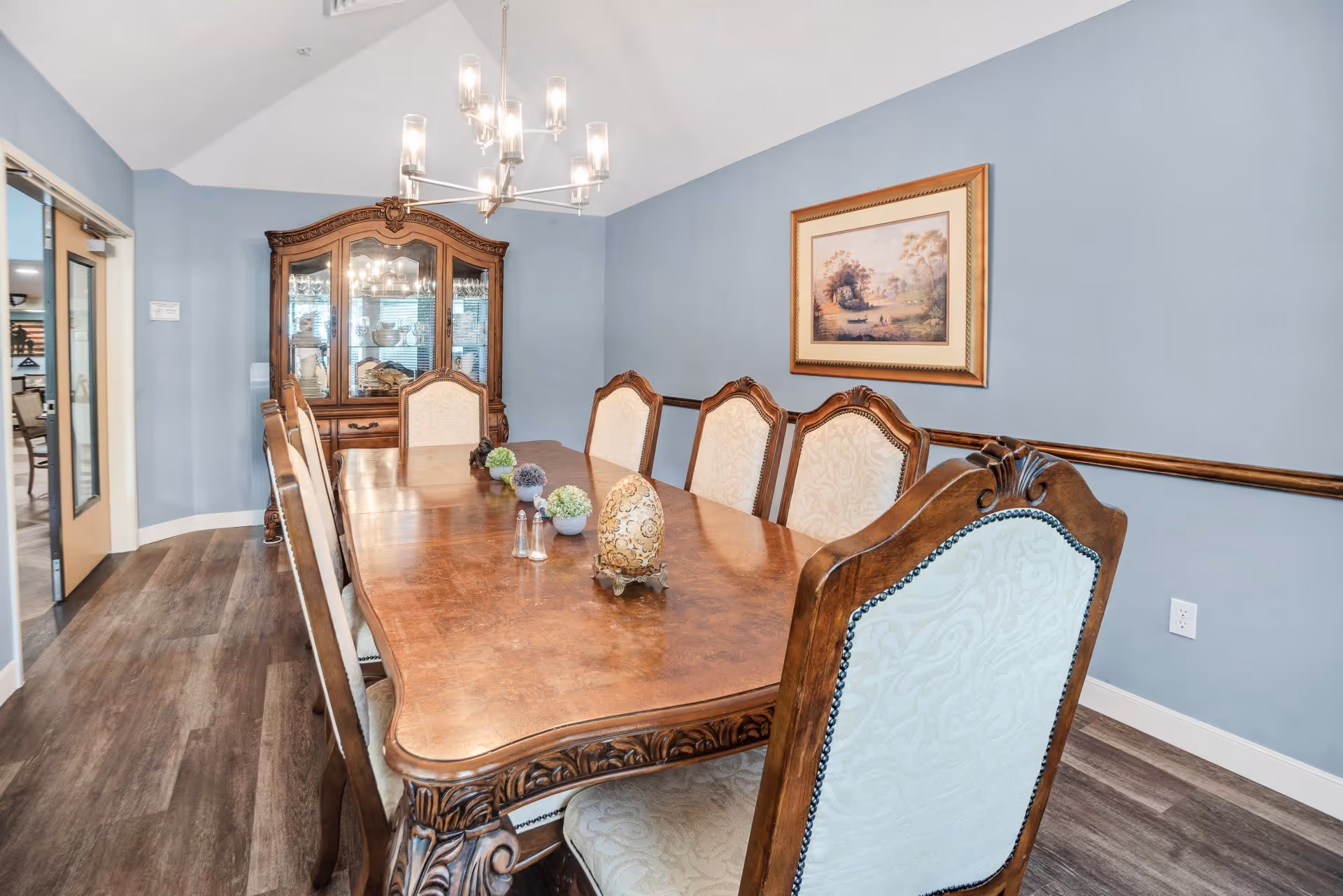A formal dining room with a long wooden table surrounded by eight upholstered chairs. The table is decorated with small ornamental items and a salt and pepper shaker set. A wooden china cabinet with glass doors is against the far wall, and a framed landscape painting hangs on the blue wall. The room has wood flooring and a modern chandelier hanging from the ceiling.