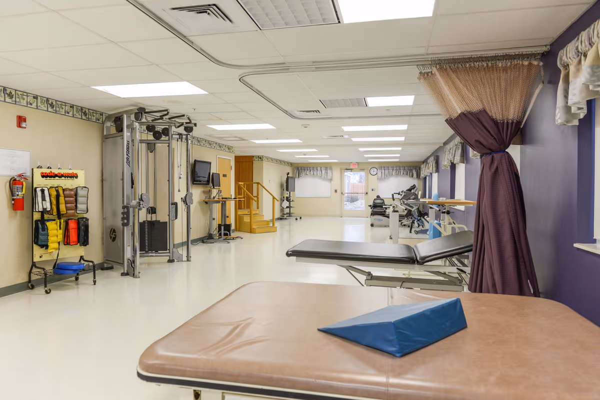A spacious rehabilitation room with exercise equipment, therapy tables, and a purple curtain tied to the side. The room has a white ceiling with fluorescent lights, beige walls with a decorative border, and a fire extinguisher mounted on the wall. There are windows with valances and a door at the far end.