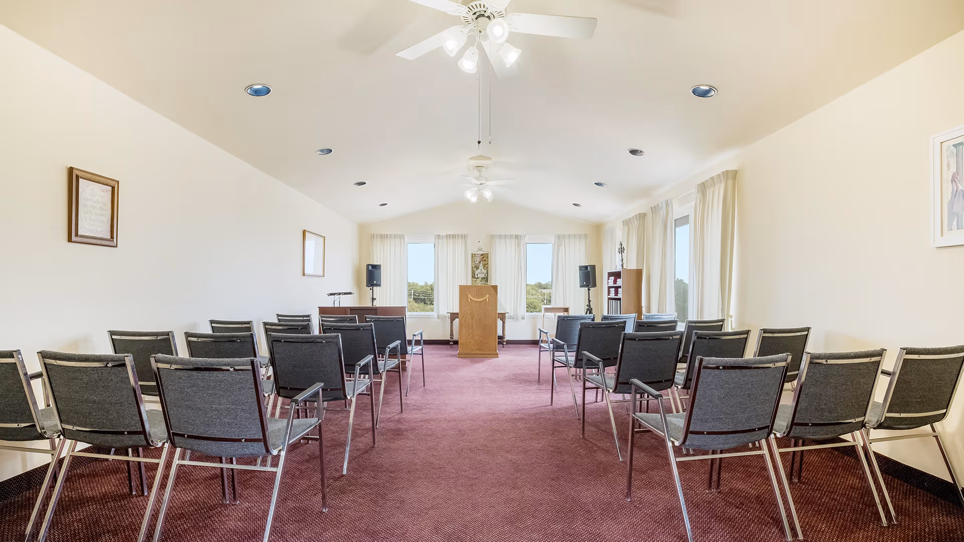 A bright room with rows of black chairs arranged facing a wooden podium at the front. The room has a maroon carpet, white walls, ceiling fans with lights, and large windows with white curtains letting in natural light. There are framed pictures on the walls and speakers positioned near the podium.