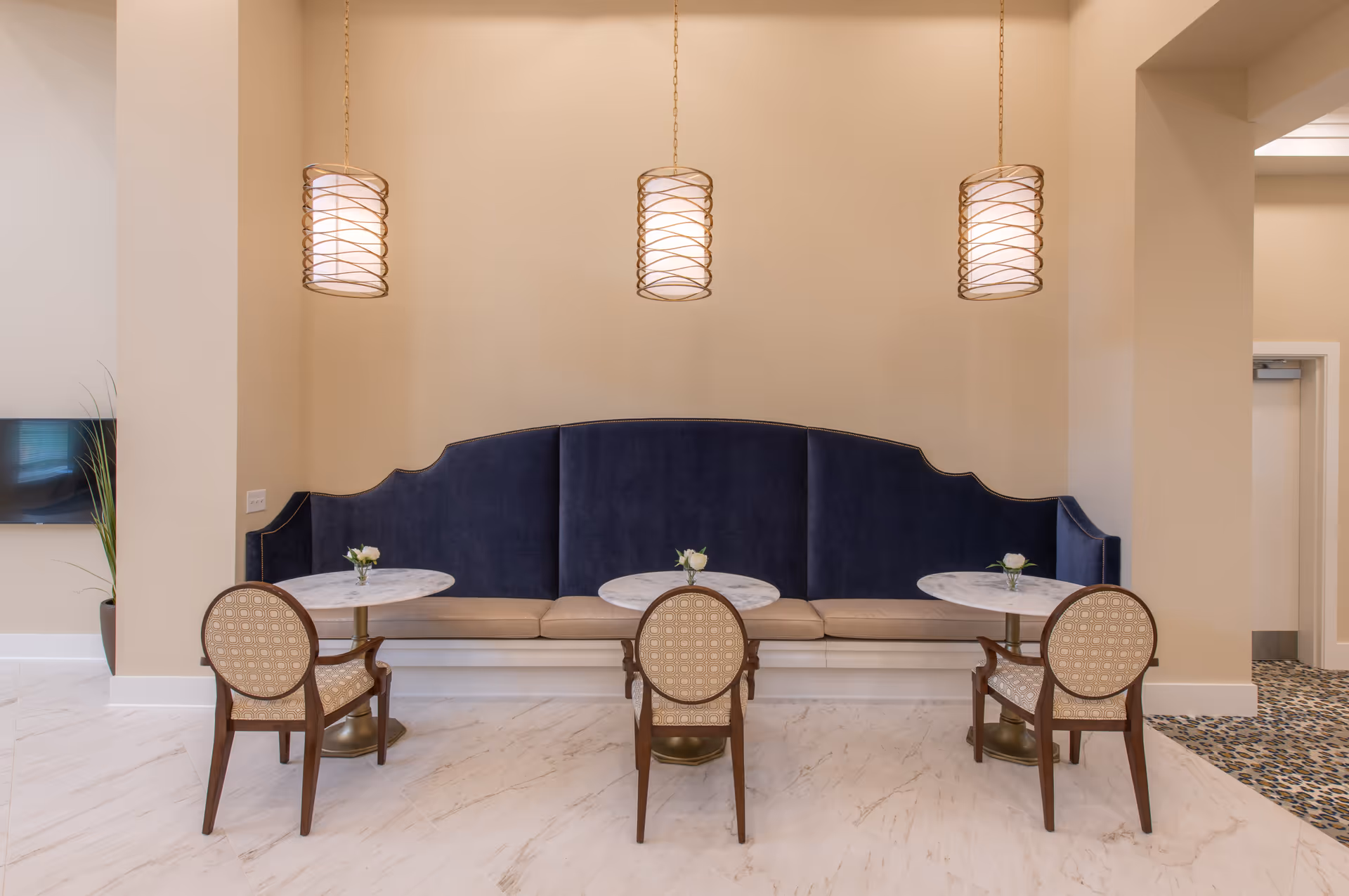 A seating area with three small round marble tables, each accompanied by a single patterned chair. Behind the tables is a long, dark blue cushioned bench with a beige seat. Above the tables hang three pendant lights with cylindrical shades and a spiral design. The floor is light-colored with a marble pattern, and the walls are painted beige.