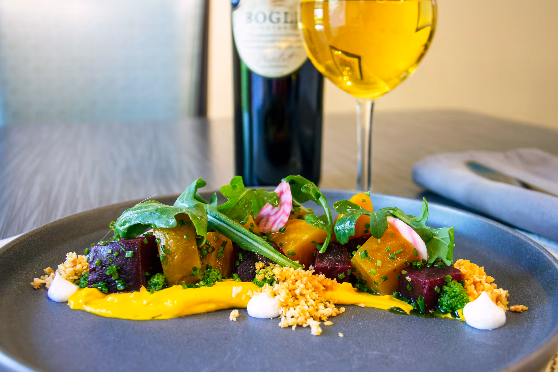 A gourmet dish featuring colorful cubes of beetroot and other vegetables garnished with fresh greens, herbs, and small dollops of sauce on a dark plate. In the background, there is a glass of white wine and a bottle of wine on a wooden table with a folded napkin and cutlery.
