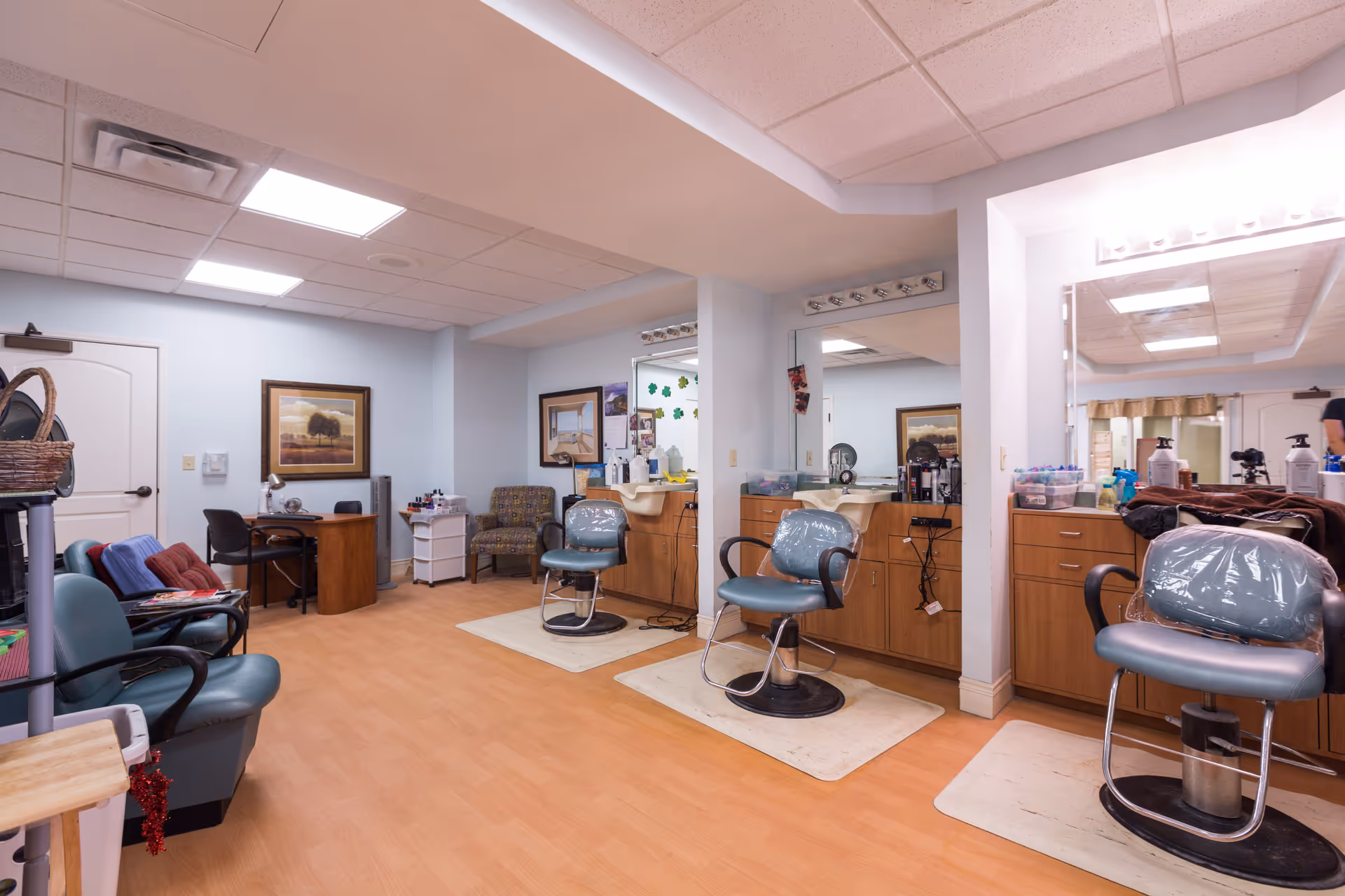 Interior view of a senior living facility's hair salon area with three salon chairs in front of mirrors and sinks, a small desk with a chair, and additional seating. The room has light blue walls, wood flooring, and ceiling lights.