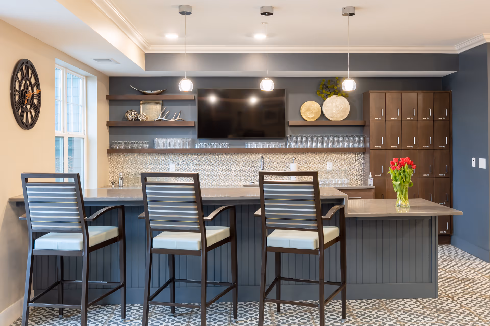 A modern kitchen area with a long island counter featuring three high chairs with striped cushions. The back wall has dark blue paint, open shelves with decorative items, a mounted flat-screen TV, and a backsplash with small mosaic tiles. There are pendant lights hanging from the ceiling and a vase with red tulips on the counter. To the right, there is a set of wooden lockers.