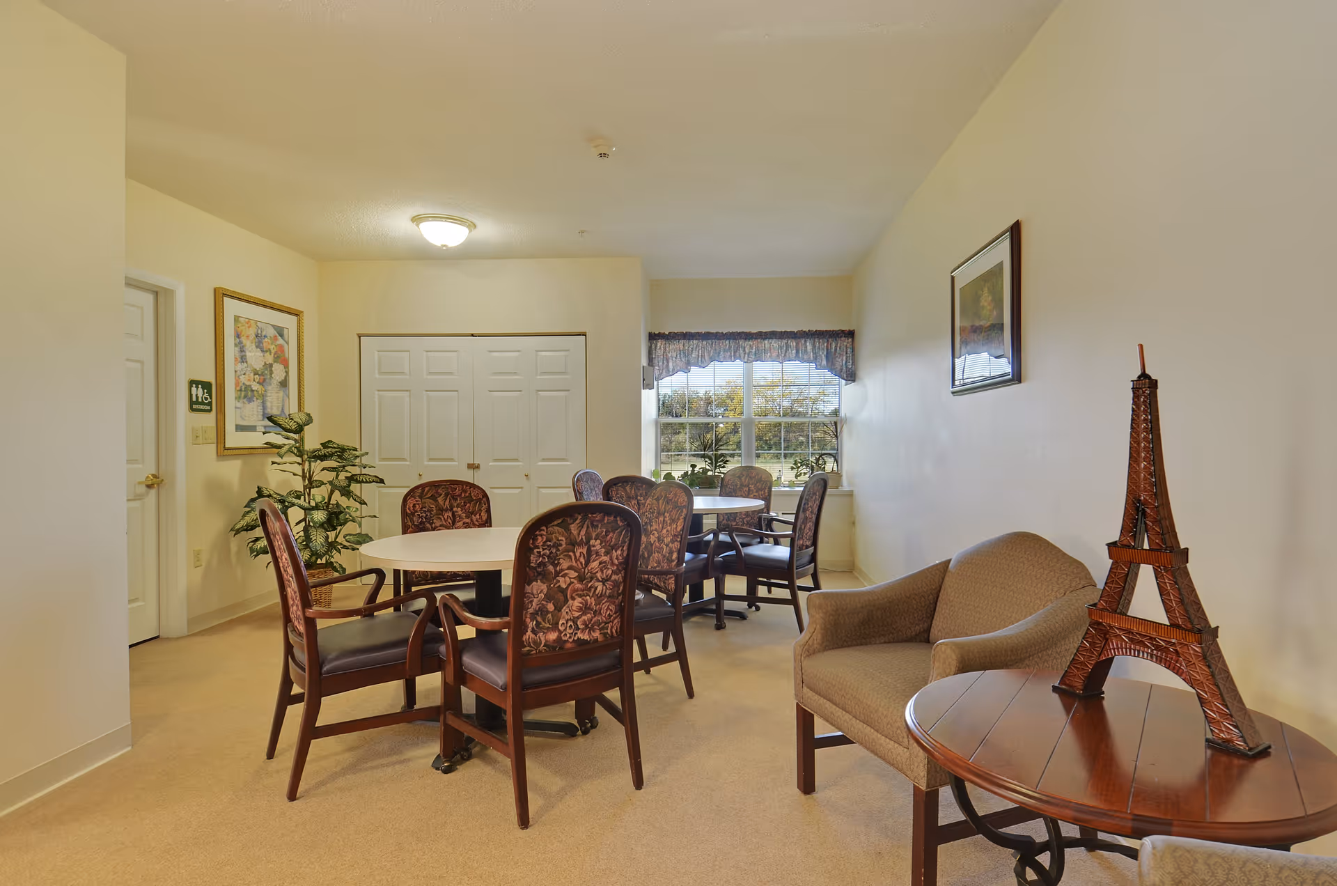 A senior living facility common area with two round tables surrounded by chairs. There is a window with a valance letting in natural light, a small armchair next to a round wooden table with a decorative Eiffel Tower model, a framed picture on the wall, and a potted plant near a door with a restroom sign.