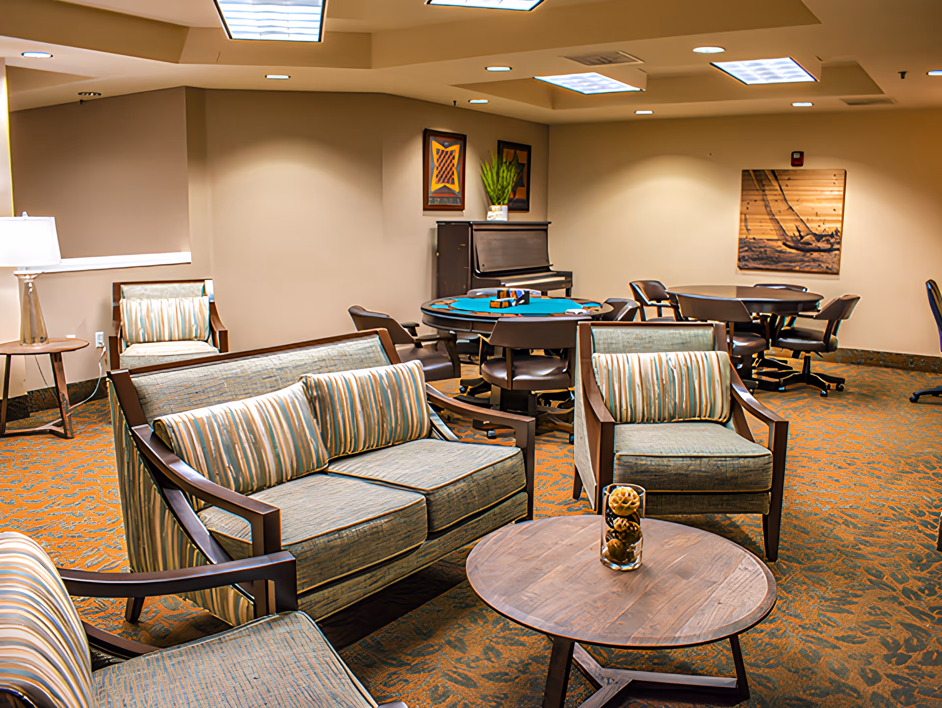 A cozy common area in a senior living facility featuring comfortable upholstered chairs and sofas with striped cushions, a round wooden coffee table with decorative items, multiple round tables with chairs, a piano against the wall, framed artwork, and warm lighting from ceiling fixtures and a floor lamp.