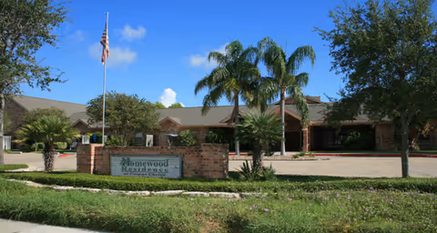 Low single-story brick senior living building with palm trees, a flagpole, and a sign on a grassy front lawn under a blue sky.