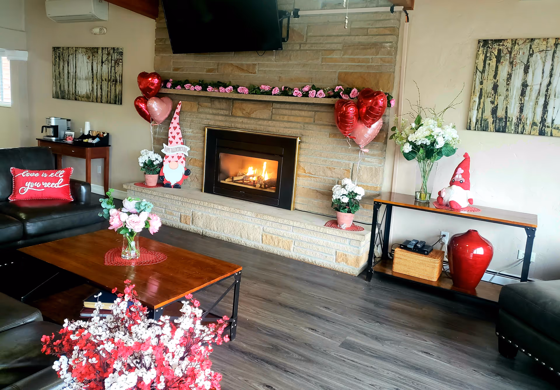Cozy living room with a stone fireplace featuring a lit fire. The mantel is decorated with pink roses and heart-shaped balloons. There are two gnome decorations on either side of the fireplace. A wooden coffee table with a vase of pink flowers sits in front of a dark leather sofa with a red pillow that reads 'love is all you need'. The room has wooden flooring and light-colored walls with two abstract paintings. A console table on the right holds a large red vase, a basket, a phone, and a vase with white flowers.