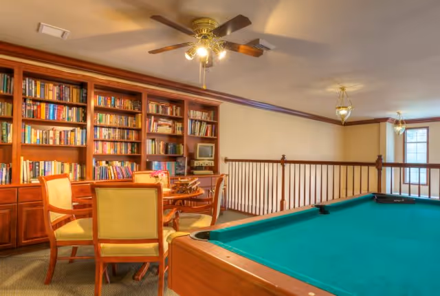 A cozy recreational room featuring a green pool table in the foreground, a wooden table with four chairs, and a large built-in wooden bookshelf filled with books and a small TV. The room has a ceiling fan with lights and two hanging light fixtures near windows with natural light.