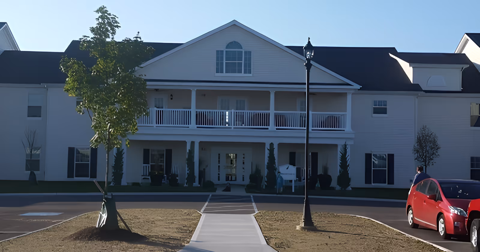 Front facade of a two-story senior living building with a central entrance, upper balcony, walkway, lamp post, and a parked red car.