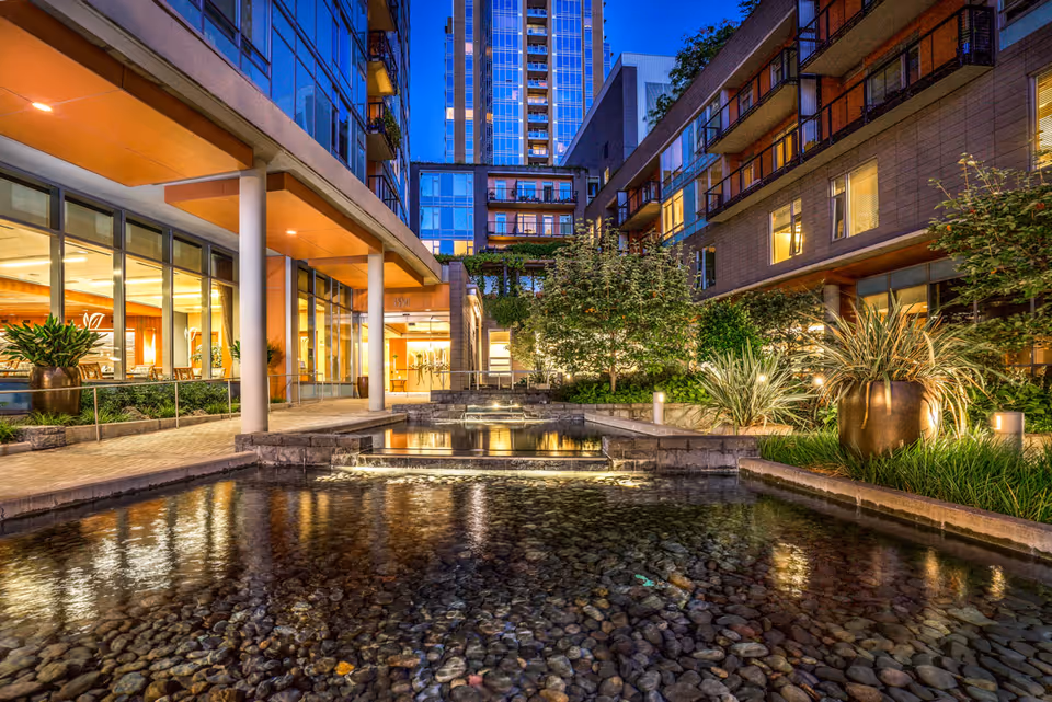 Evening view of a modern senior living facility courtyard with a shallow water feature filled with stones, surrounded by plants and illuminated by soft lighting. The building has large windows and balconies, with a tall city building visible in the background.