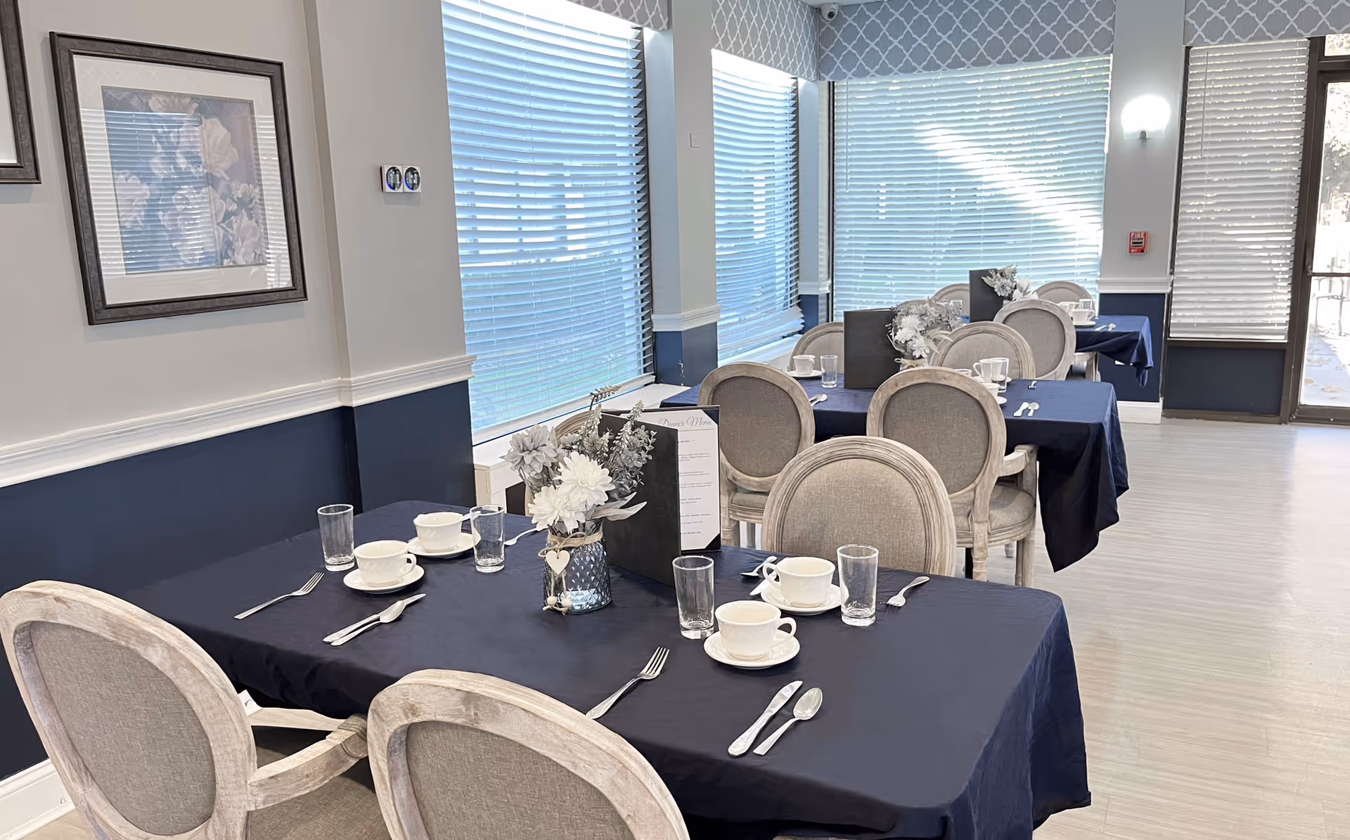Dining room with navy tablecloths, set place settings, floral centerpieces, and upholstered chairs by large windows.