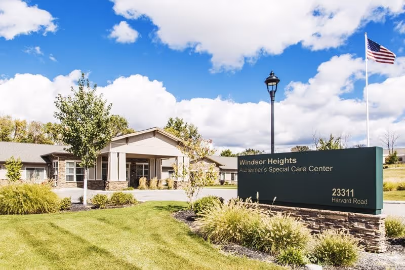 Exterior view of Windsor Heights assisted living building with an entrance canopy, landscaped lawn, and a large sign showing the facility name and address under a blue sky.