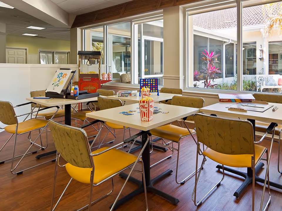 Bright communal activity/dining room with tables and yellow chairs, board games and a popcorn machine by large windows.
