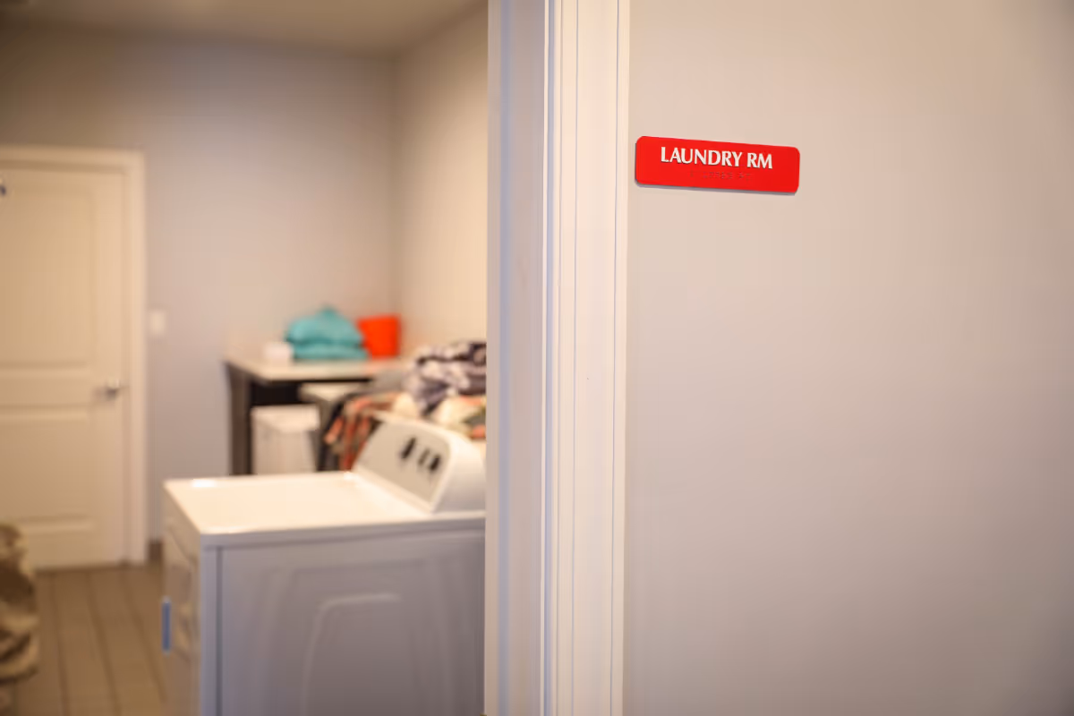View of a laundry room with a washing machine in the foreground and folded clothes on a counter in the background. A red sign on the wall outside the room reads 'LAUNDRY RM'.