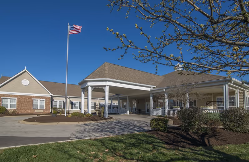Exterior view of Crescent Place Assisted Living facility showing a covered entrance with white pillars, a circular driveway, an American flag on a flagpole, and landscaped bushes and trees under a clear blue sky.