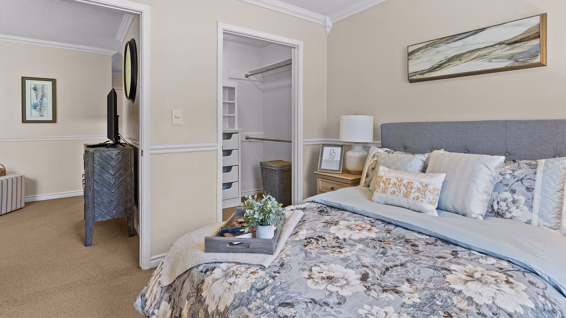 A cozy bedroom with a bed featuring floral bedding and multiple pillows. A wooden nightstand with a lamp and framed picture is beside the bed. There is an open walk-in closet with shelves and hanging rods. A TV on a wooden stand is visible in the adjacent room, along with a framed picture on the wall.