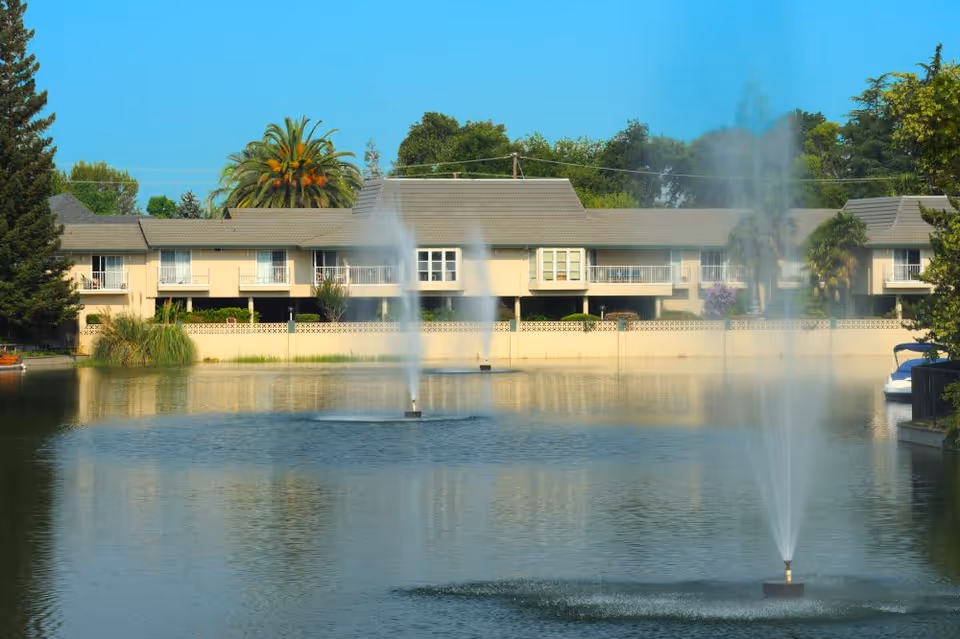 View of a senior living facility building behind a large pond with three water fountains spraying water upwards, surrounded by trees and clear blue sky.