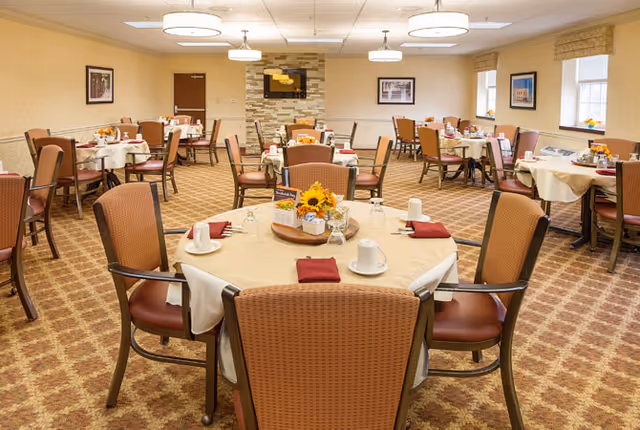 A spacious dining room at Leighton Place featuring round tables set for meals, with chairs and decorative centerpieces including sunflowers. The room has a warm color scheme and is well-lit with overhead lighting.