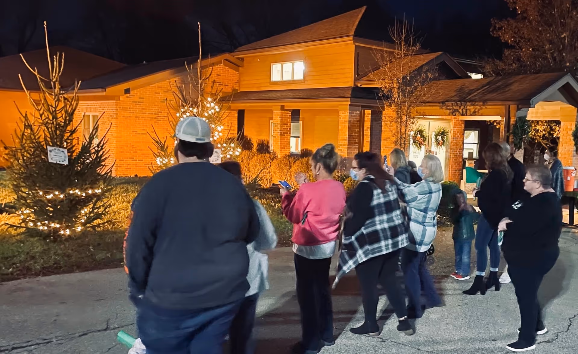 A group of people gathered outside the lit brick front of a senior living facility at night, with decorated evergreen trees and wreaths.