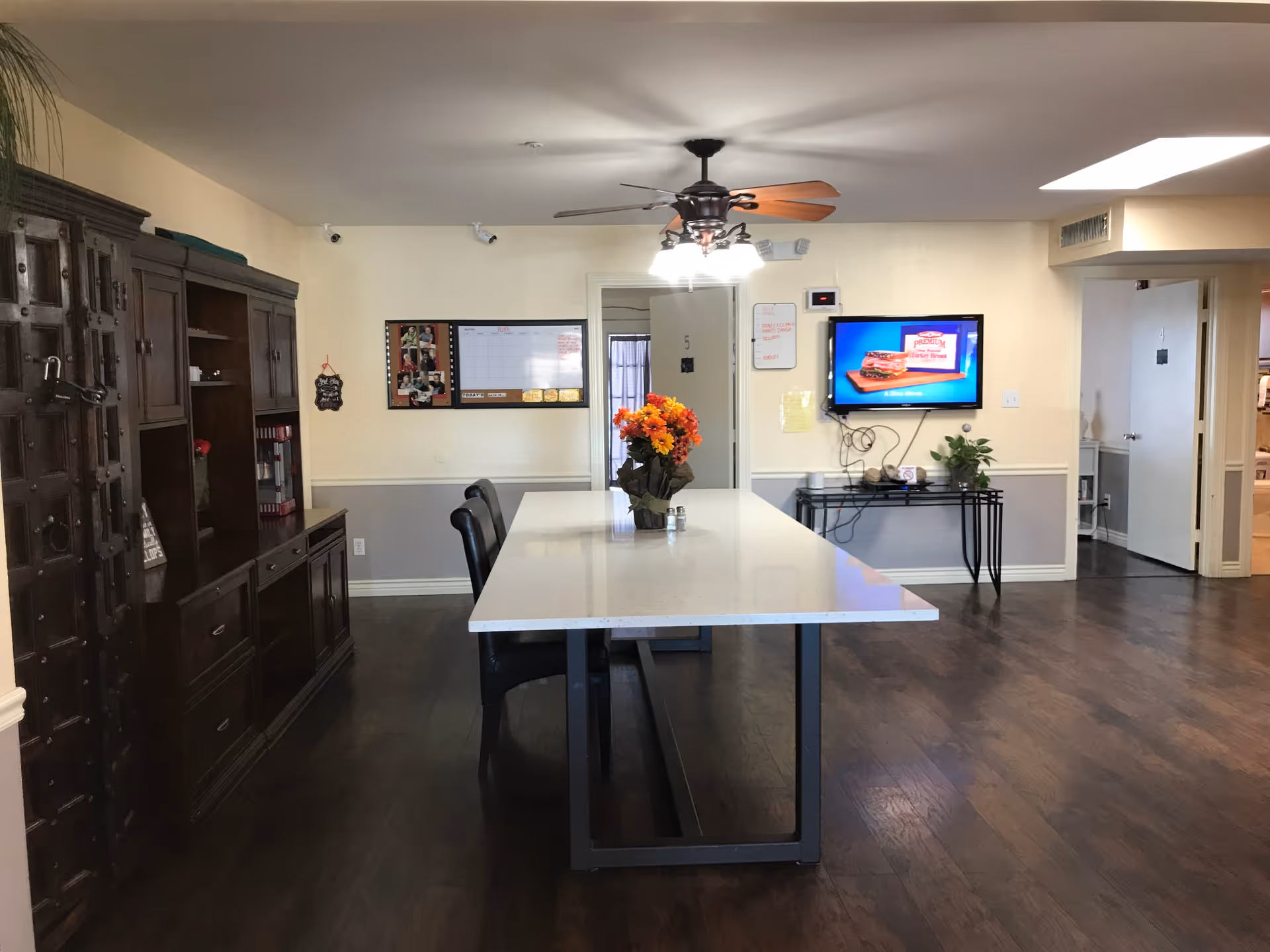 Interior view of a communal room with a long white table and black chairs around it. A vase with orange and yellow flowers is placed on the table. On the left side, there is a large dark wooden cabinet. The walls are light-colored with a white chair rail. A ceiling fan with lights hangs above the table. On the far wall, there is a flat-screen TV mounted, displaying an advertisement, and a bulletin board with notes and photos. Doors labeled with numbers are visible in the background.