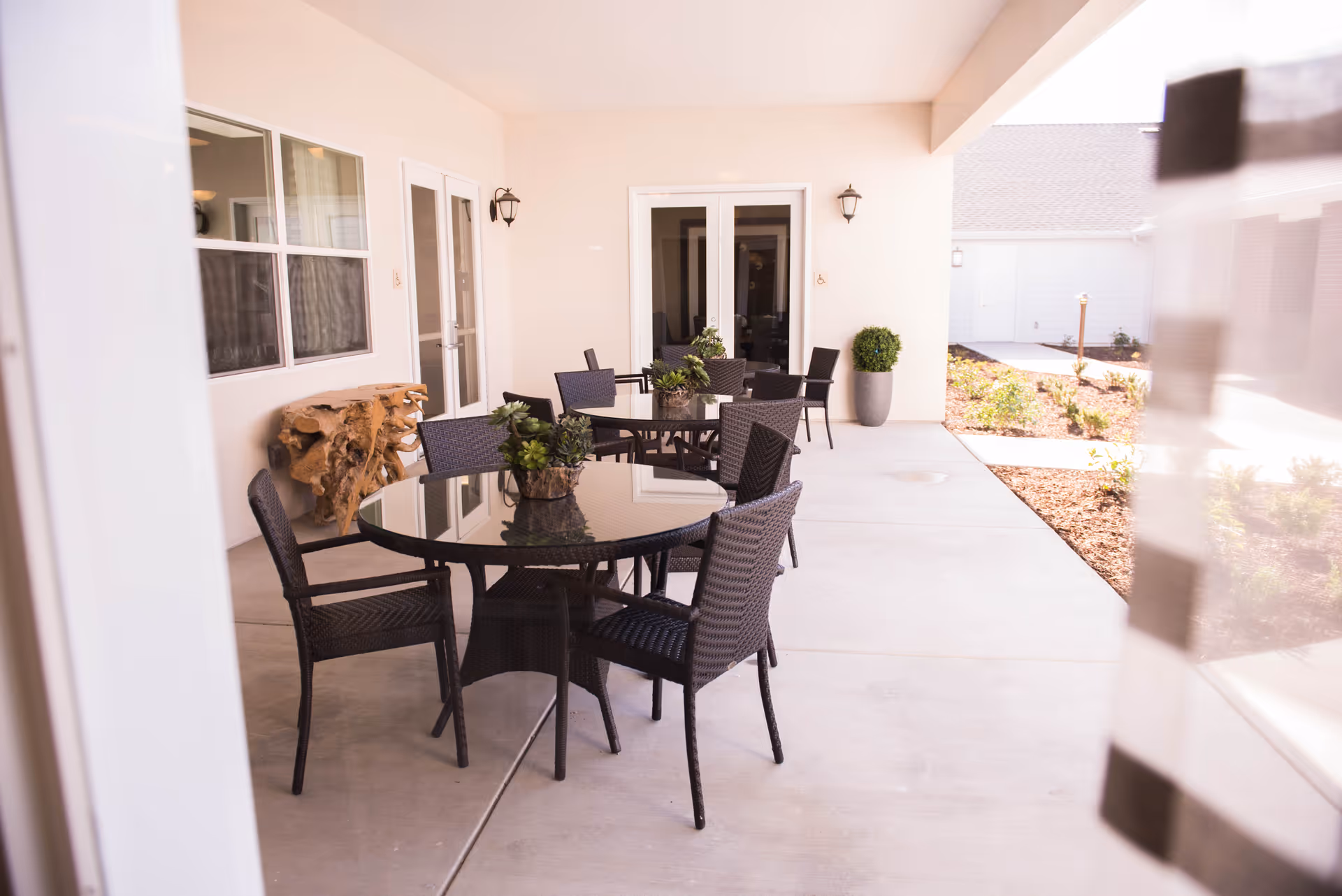 Outdoor covered patio area with two round glass-top tables surrounded by black wicker chairs. Each table has a small potted plant centerpiece. The patio is adjacent to a building with beige walls, white-framed windows, and double glass doors. There are wall-mounted lantern-style lights and a large potted plant near the doors. The patio overlooks a landscaped area with mulch and small plants.