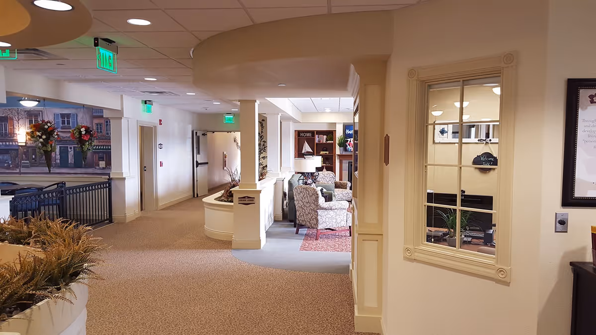 Carpeted interior hallway of a retirement community leading to a small lounge with upholstered chairs, plants, and decorative wall window.