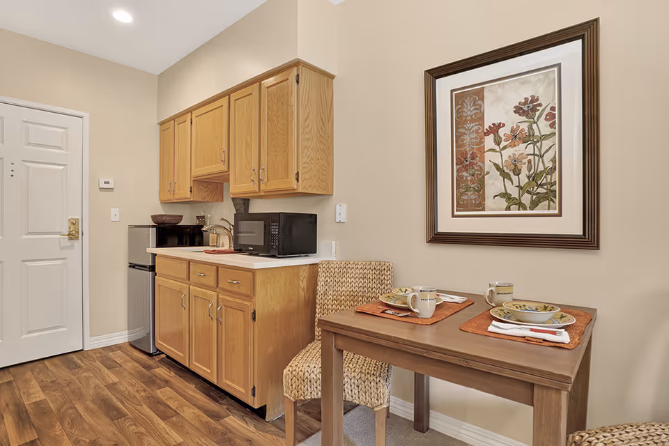A small kitchen area with wooden cabinets, a microwave, and a mini refrigerator. Next to the kitchen is a wooden dining table set for two with plates, bowls, mugs, and utensils on orange placemats. A framed floral artwork hangs on the beige wall above the table. The floor is wood, and there is a white door in the background.