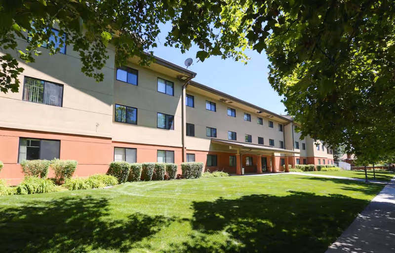 Senior living facility exterior with manicured lawn, trees, and a welcoming entrance.