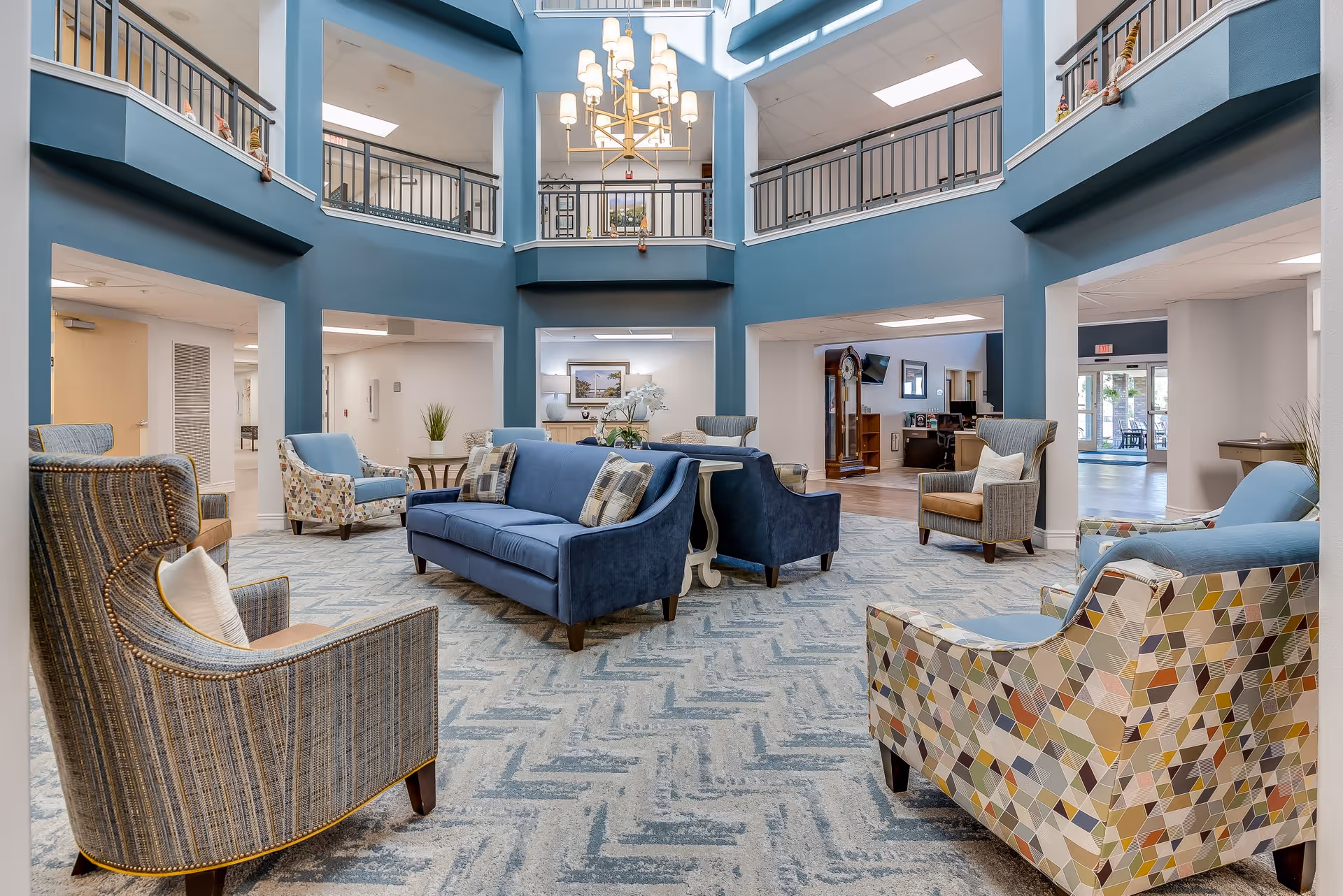 A spacious and well-lit senior living facility common area with blue walls and a high ceiling. The room features a variety of comfortable seating options including blue sofas, patterned armchairs, and striped chairs arranged around small tables. A large chandelier hangs from the ceiling, and there are railings on the second floor overlooking the area. The space has a welcoming and cozy atmosphere with decorative plants and framed artwork on the walls.