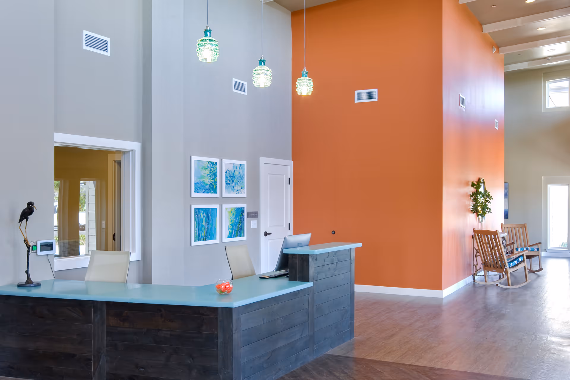Reception area with a modern desk featuring a glass countertop, two chairs, and a computer. The walls are painted gray and orange, with four abstract blue and green paintings on the gray wall. Three pendant lights hang from the ceiling. In the background, there are two wooden rocking chairs with cushions and a small plant on the orange wall.