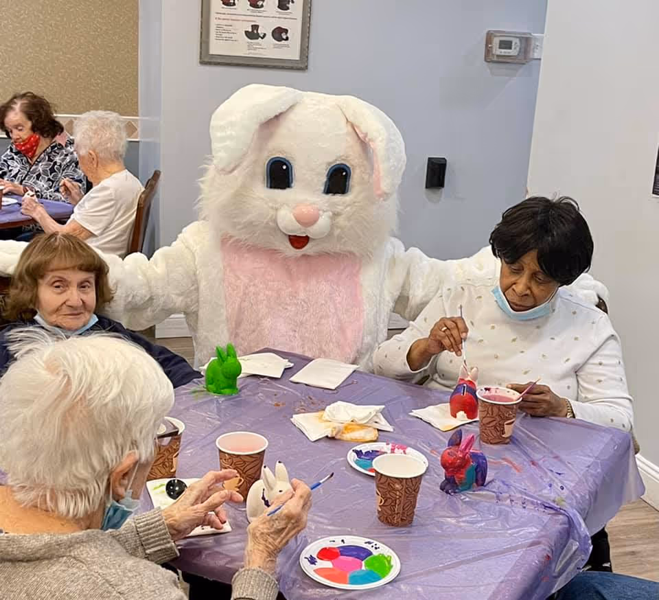 Seniors seated around a table painting ceramic bunnies while a person in a large white Easter bunny costume stands behind them.