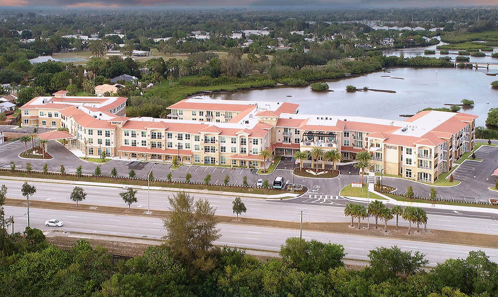 Aerial view of Heartis Venice Assisted Living facility showing a large, three-story building with a red-tiled roof surrounded by parking lots, palm trees, and greenery. The building is situated near a body of water with a scenic landscape in the background.