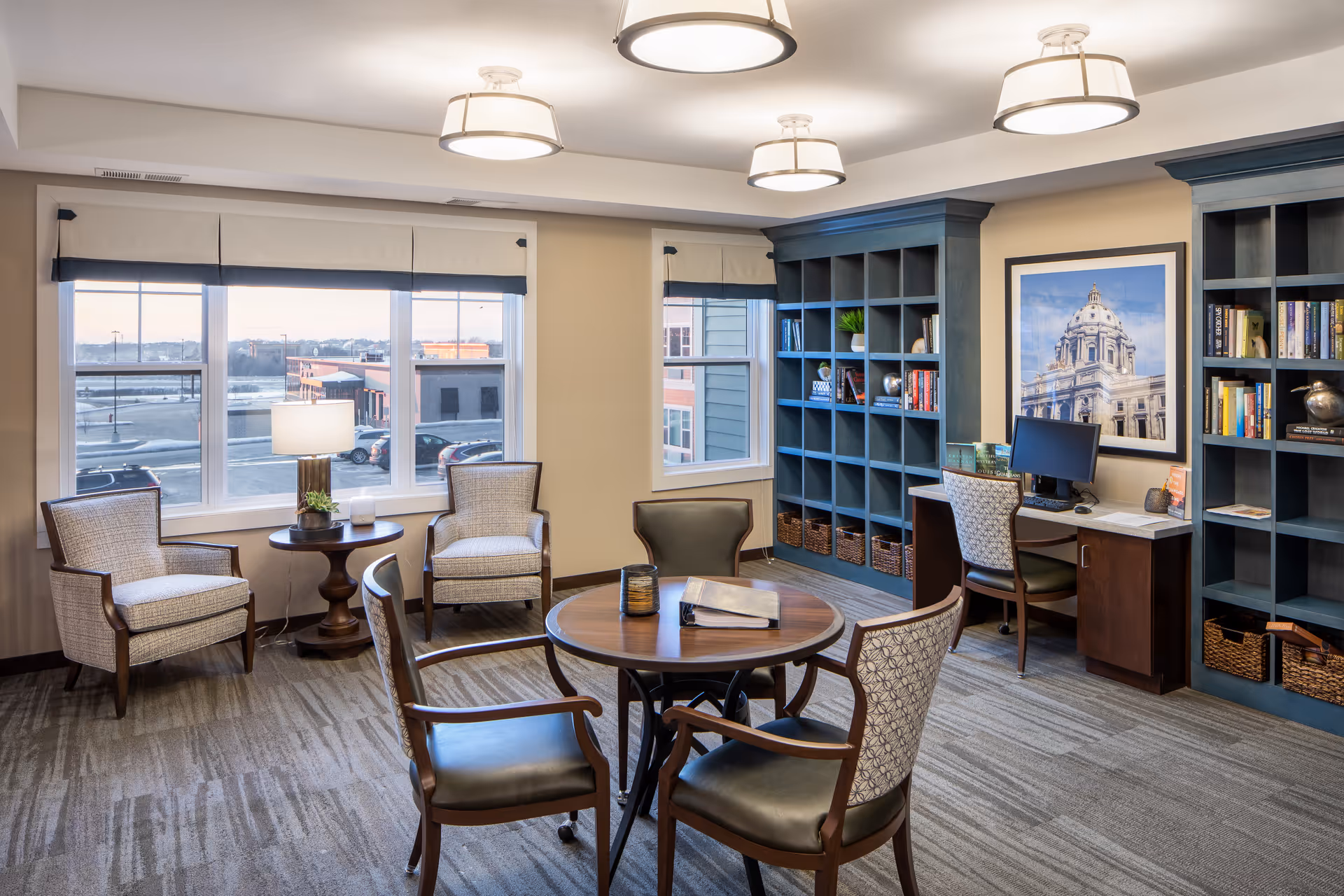 Well-lit community lounge with a round table and chairs, armchairs by large windows, built-in bookshelves and a computer desk.