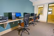A small computer room with three desktop computers on a long wooden desk against a green wall. Each computer has a black office chair in front of it. The room has beige carpet flooring and a doorway leading to another area with yellow walls.