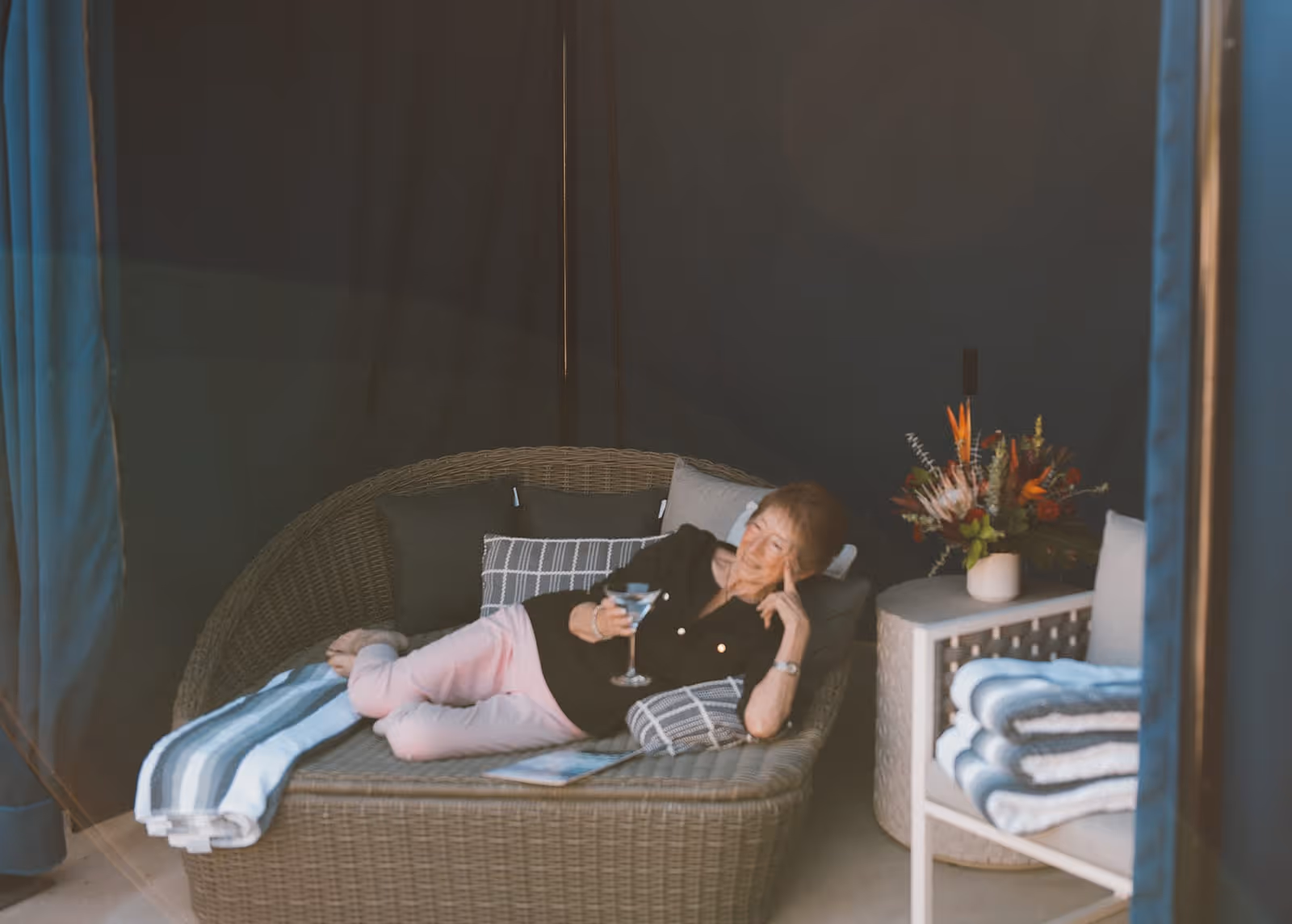 An elderly woman relaxing on a wicker lounge chair with cushions, holding a drink in her hand. She is indoors with dark curtains in the background, next to a small table with a vase of flowers and folded towels on a nearby chair.