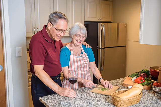 An older couple in a kitchen preparing food, with the woman chopping herbs at a counter and the man standing beside her holding a glass of wine.