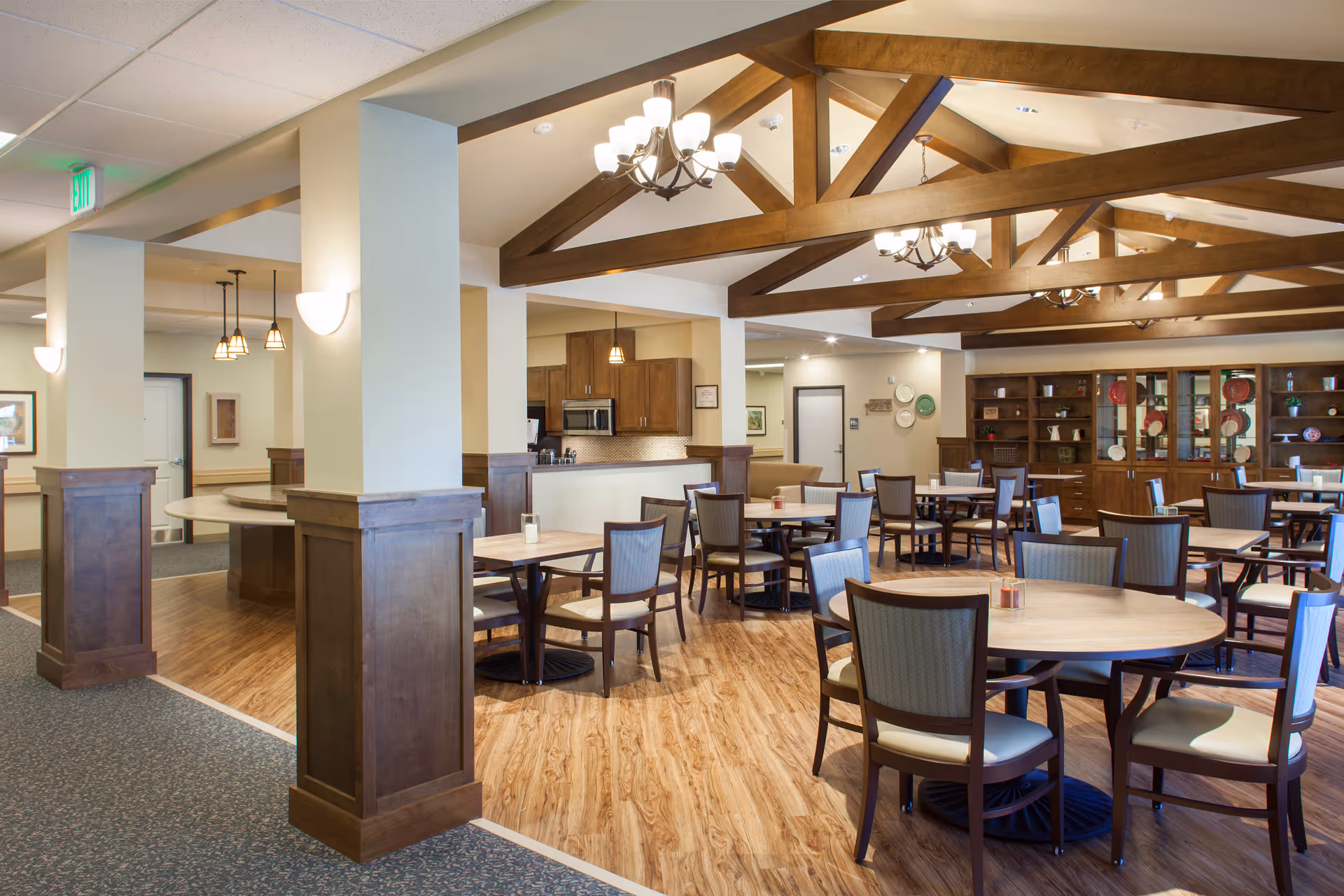 A spacious dining area in a senior living facility featuring multiple round and square wooden tables with cushioned chairs. The room has wooden beams on the ceiling with chandeliers, a wooden floor section, and a carpeted area. There is a kitchen area with cabinets and a microwave in the background, and a large wooden cabinet with decorative plates and items on the right side.