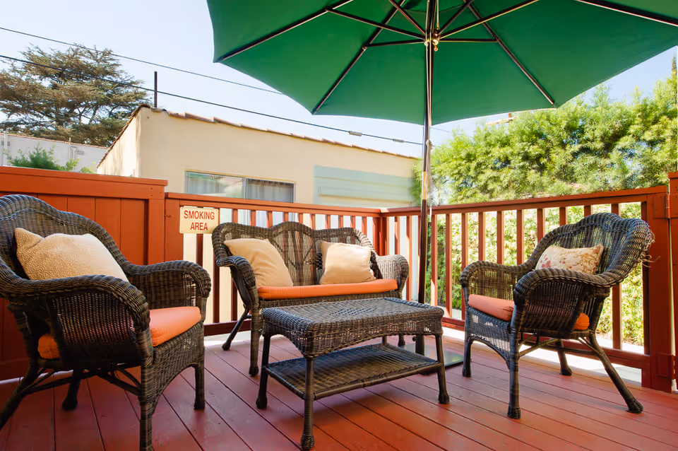Outdoor patio area with wicker furniture including two armchairs and a loveseat with orange cushions and pillows, a wicker coffee table, and a large green umbrella providing shade. The patio has red wooden flooring and railing, with greenery and a building visible in the background. A small sign on the railing reads 'SMOKING AREA'.