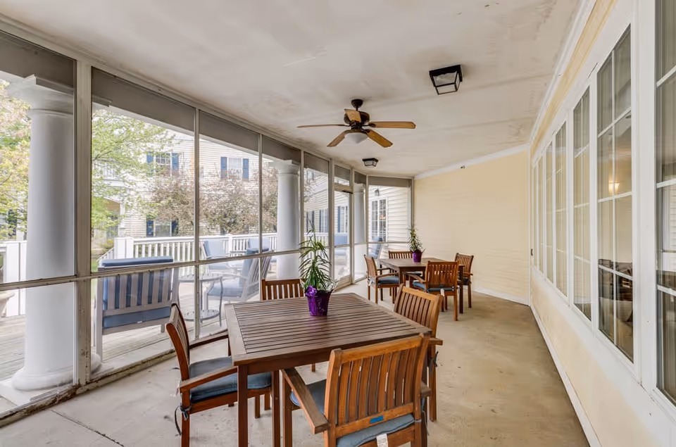 Screened covered patio with wooden tables and chairs, a ceiling fan, and potted plants overlooking an outdoor deck.