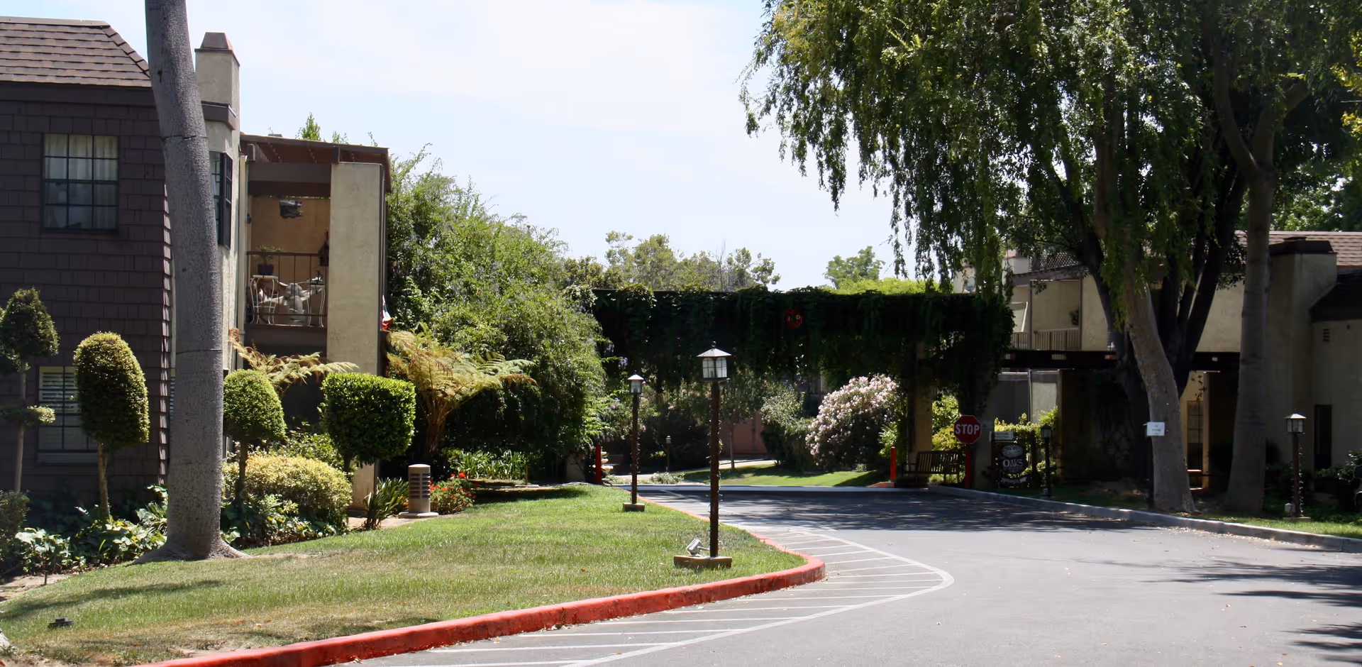 Outdoor view of a senior living facility with a paved driveway, manicured lawns, trimmed bushes, tall trees, and residential buildings on either side. There is a stop sign and a covered entrance with greenery hanging over it in the background.